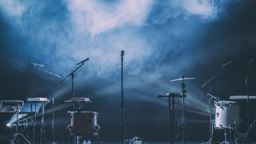 Concert stage setup with microphones, drums, keyboards, and lighting equipment under a smoky blue backdrop