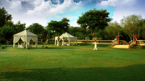 Poolside Lawn at Clarks Khajuraho featuring two event canopies set up on a spacious green lawn surrounded by lush trees, garden fences, and a bright, cloudy sky.