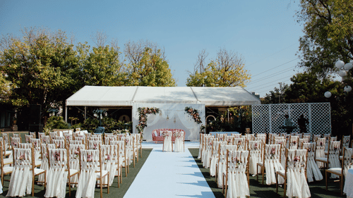 Elegant outdoor wedding venue featuring a white aisle, floral wedding arch, beautifully decorated guest seating, and a stylish stage set amidst lush greenery under a clear blue sky.