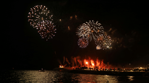 Fireworks explode in the night sky above a body of water reflecting the lights of a nearby dock or shore.