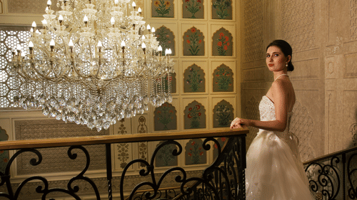 A woman in a white gown poses by a staircase beneath a grand crystal chandelier and floral wall motifs -  Heritage Village Resorts & Spa, Manesar