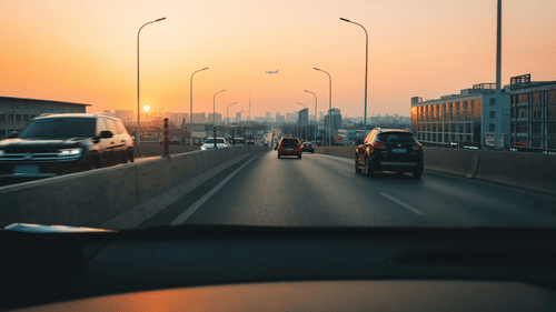 A view from inside a car showing a road with multiple vehicles, streetlights, buildings on both sides, and the sky during sunset.