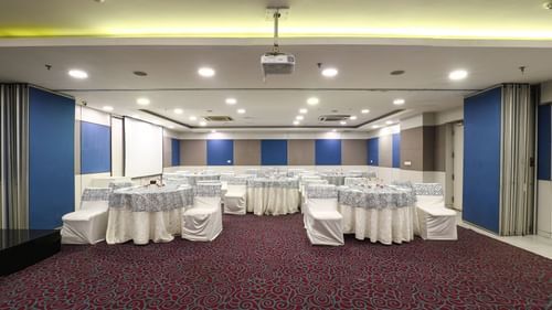 A large and well-lit conference hall with round tables, white chair covers, and a modern projector setup, perfect for professional events and meetings.