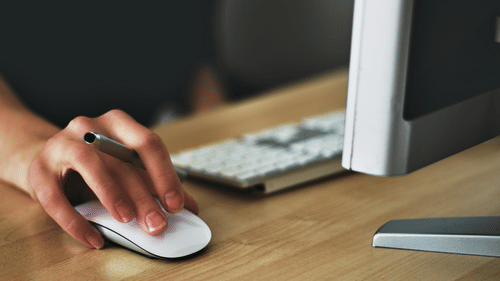 A person using a computer mouse at a desk, with a monitor and keyboard placed beside it.