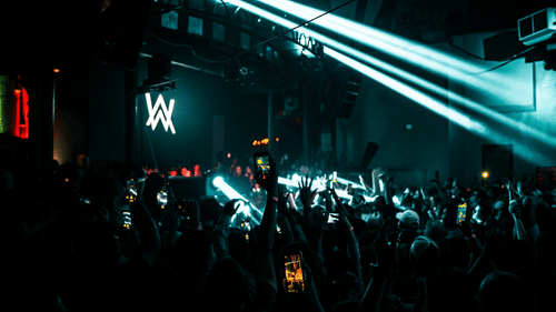  A packed nightclub with a large crowd facing a stage with a prominent AW logo. Multiple spotlights illuminate the scene, and the audience is holding up their phones, capturing the moment.