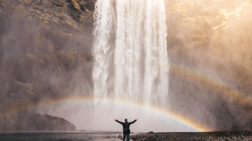 A person standing at the edge looking at the waterfalls with a rainbow in front of the falls