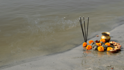 A picture of lit incense sticks, flowers and a copper bowl next to a river