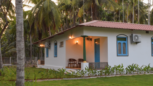 A white resort cottage with a red roof and blue window frames, surrounded by palm trees and green lawns - Ibex Resorts, Coimbatore (Kakarla)
