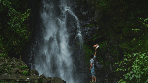 A person with a rucksack stands before a tall, misty waterfall cascading down a rocky cliff in a lush green forest.