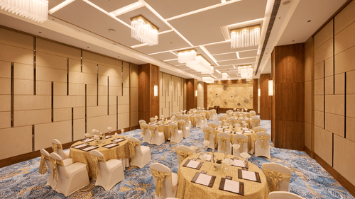 A formal event space is prepared with round tables covered in gold cloths, each with place settings and surrounded by chairs adorned with gold sashes, under a modern ceiling light design. - Iris Sarovar Portico, Sector 85, Gurugram
