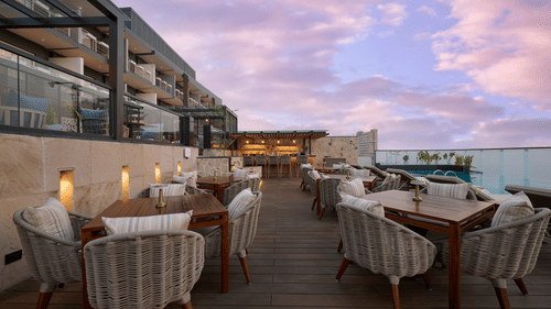 An outdoor dining area at a hotel overlooks a swimming pool, with tables and chairs featuring plush cushions, set against a backdrop of a colourful evening sky. - Iris Sarovar Portico, Sector 85, Gurugram