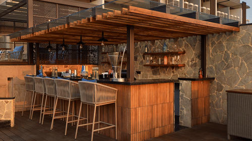 A stylish outdoor bar area with a wooden countertop and stools is set up, featuring shelves stocked with bottles and glasses, ready to serve drinks in a relaxed setting. - Iris Sarovar Portico, Sector 85, Gurugram
