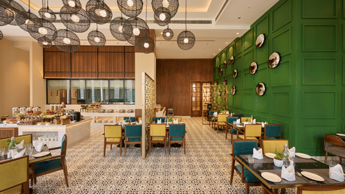 A stylish restaurant space features a buffet area, dining tables with colourful chairs, a patterned floor, and unique spherical pendant lights hanging from the ceiling. - Iris Sarovar Portico, Sector 85, Gurugram