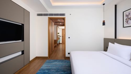 A modern hotel bedroom features a large bed with white linens, a blue rug on the wooden floor, a wall-mounted television, and an open doorway leading to another part of the suite. - Iris Sarovar Portico, Sector 85, Gurugram