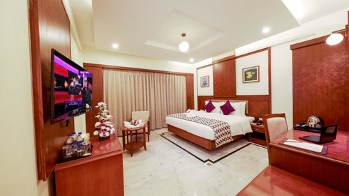 Club Room at JP Hotel in Chennai featuring a plush bed with wall hangings over it on the two sides of the wall facing a seating space by the closed window and a TV on the wall facing the study desk