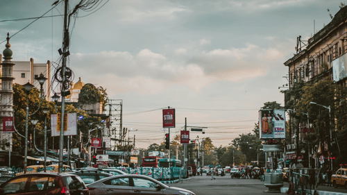 A busy city street scene is shown with cars, trams, and buildings under a cloudy sky.
