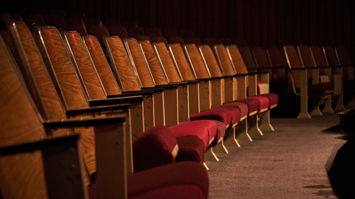 Row of empty vintage-style theatre seats with red cushions and wooden backs, set in a dimly lit auditorium.