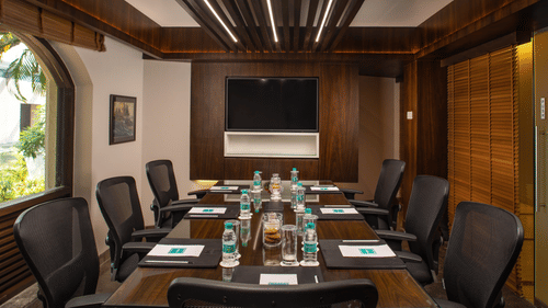 A view of the Boardroom meeting hall at Kenilworth Resort and Spa, featuring comfortable chairs, a large desk, a tv, books and pens on the table with water bottles. 