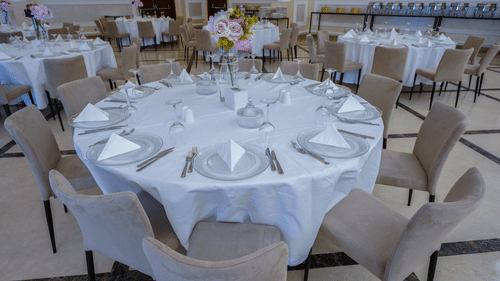 A banquet hall with round tables set for an event. Each table is covered with a white tablecloth and arranged with plates, napkins, and cutlery. Neutral-coloured chairs surround the tables, and a floral centrepiece adorns each at La Maison Hotel, Doha.