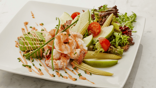 A shrimp salad on a white plate with lettuce, sliced avocados, and a lime wedge. The plate is garnished with a drizzle of sauce.