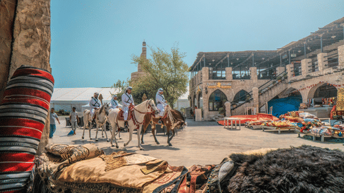 A vibrant marketplace scene in Doha, Qatar, features individuals on horseback in traditional attire, with various textiles and goods displayed in the foreground and traditional architecture in the background.
