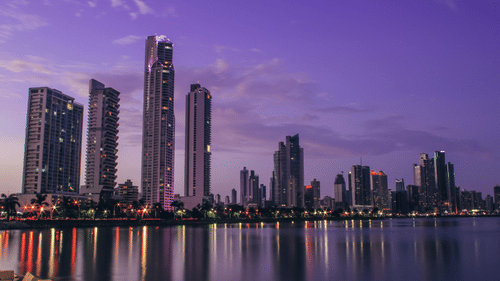 A purple-hued sunset over a city skyline with tall buildings reflected in the calm water below.