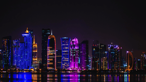 The illuminated skyline of Doha's Dhow Harbour at night, with vibrant lights from the skyscrapers reflecting on the water.