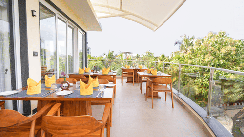 A bright outdoor Abobe Lounge with wooden tables and chairs on a balcony overlooking greenery at MGM Beach Resorts, ECR Chennai.