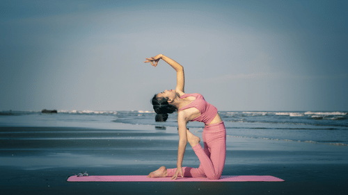A woman performing a yoga pose on the beach with a pink mat placed below her.