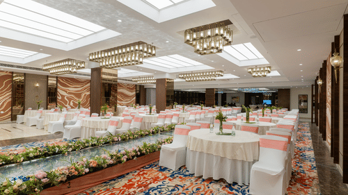An event space at Nirvana Sarovar Portico, Gorakhpur featuring round tables, dressed chairs, floral arrangements, and ceiling lights.