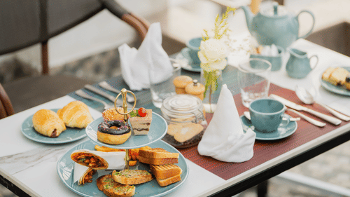 An outdoor table setting at Nirvana Sarovar Portico, Gorakhpur featuring a teapot, teacups, pastries, and a 2 tier tray of snacks.  