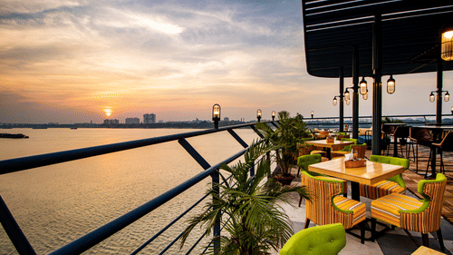 A scenic rooftop restaurant with colorful chairs, wooden tables, and a view of the sunset over a river. - Polo Floatel