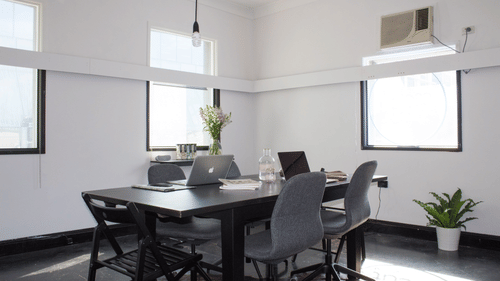 An office-style meeting room with walls, a large dark table, and several chairs. The room has three windows and a small potted plant in the corner.