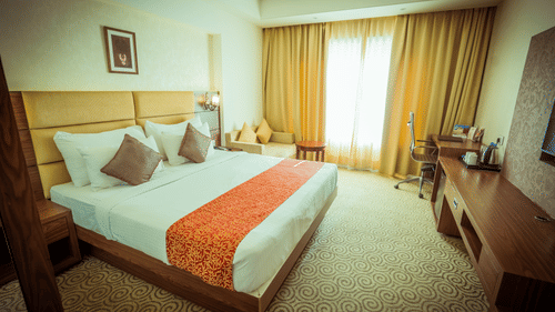 A spacious room at Pride Hotel and Convention Centre, Indore with a large bed, wooden furniture, a work desk, and a window with both sheer and dark curtains.