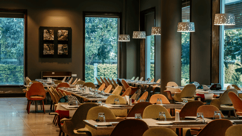 Dining area of a restaurant at a property by Pride Group of Hotels featuring rows of neatly arranged chairs and tables, large glass windows and modern lighting.