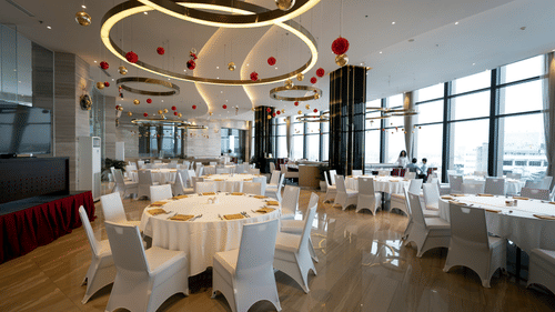 A bright, modern event hall or restaurant space set up with round tables, white chairs, and elegant ceiling lights, with large windows overlooking a city.