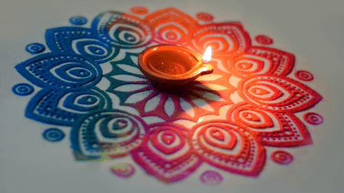 a rangoli made by hand with different colours and a diya lit up in the centre of the rangoli