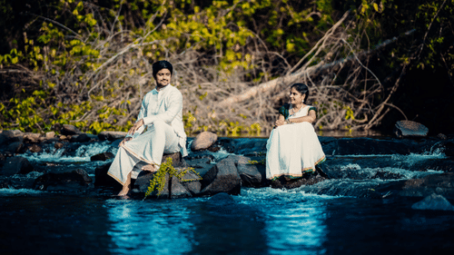a bride and groom posing while sitting on different rocks in a waterbody wearing the traditional wedding attire