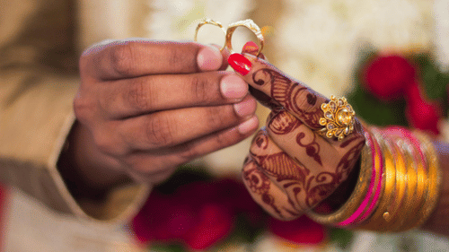 A couple exchanges wedding rings during a traditional Indian wedding ceremony, showcasing the groom's hand holding a gold ring and the bride's hand, adorned with henna and bangles, holding a diamond ring.