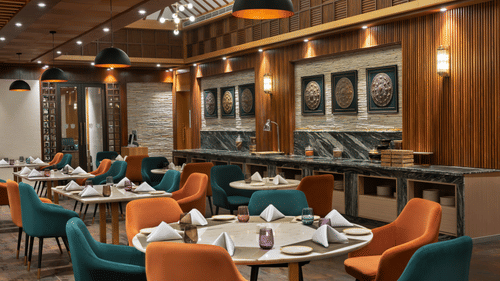 Guras restaurant interior at Royal Tulip Shivapuri National Park - Kathmandu featuring round tables, orange and teal chairs, and wood panelling.