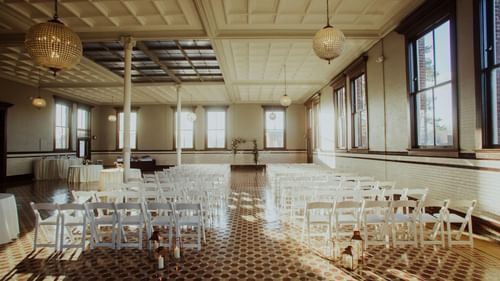 A large, empty hall with many windows and rows of white chairs and tables set up for an event.