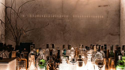A dimly lit bar shows a counter with illuminated bottles and glasses, set against a textured wall with a faint inscription and bare branches.