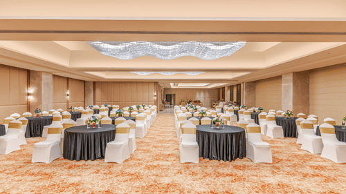 A wide view of a ballroom at Sarovar Premiere Alwar with round tables and chairs arranged on a patterned carpet floor.