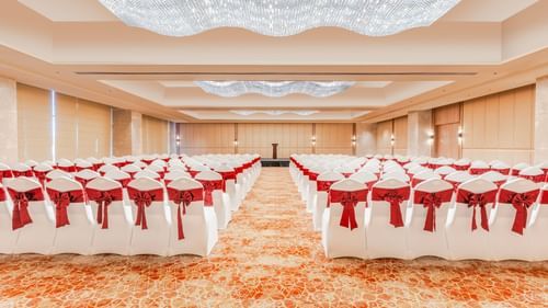 A banquet hall at Sarovar Premiere Alwar with rows of chairs facing a podium on a stage with a chandelier.