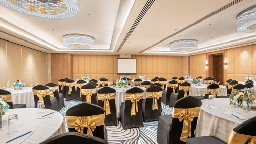 A conference room at Sarovar Premiere Alwar with round tables and chairs with gold sashes and a projector screen.