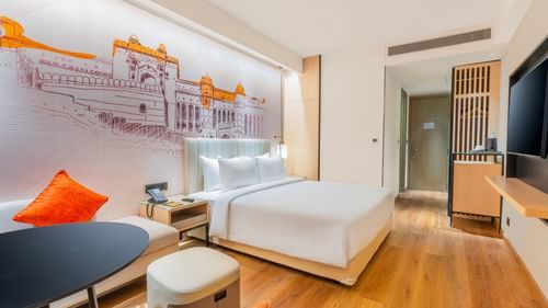 A view of the interior of a room at Sarovar Premiere Alwar, showing a bed, a side table, a chair, and a mural on the wall.