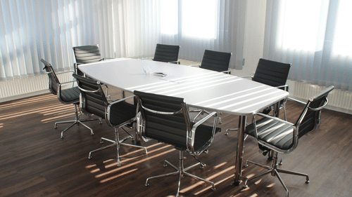 A bright, empty modern boardroom with a large white oval table and black rolling office chairs.