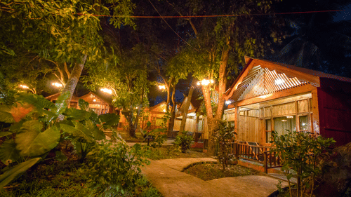Facade view of a row of wooden cottages during night time with the lights on and greenery next to it - Symphony palms beach resort and spa.