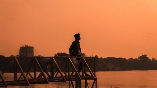 a person standing on a pier looking at the sunset. A waterbody and trees also in the image