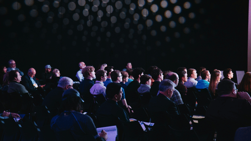 Audience seated in dimly lit room, listening attentively. A backdrop of circular lights adds ambiance. The mood is focused and engaged.